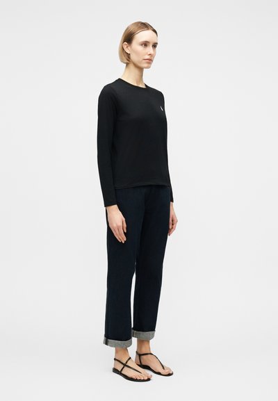 Γυναίκα στέκεται, φοράει μαύρο μακρυμάνικο πουκάμισο, σκούρο παντελόνι με ρέλι και μαύρα σανδάλια με λουριά και επίπεδη σόλα, μπροστά σε ένα απλό ανοιχτό φόντο.