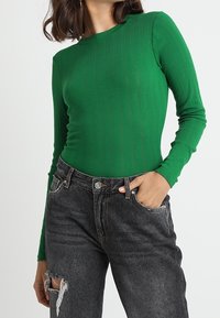 Grünes geripptes Langarmoberteil mit Rundhalsausschnitt. Kombiniert mit hochgeschnittenen, schwarzen, distressed Jeans mit auffälligen Rissen und Taschen.
