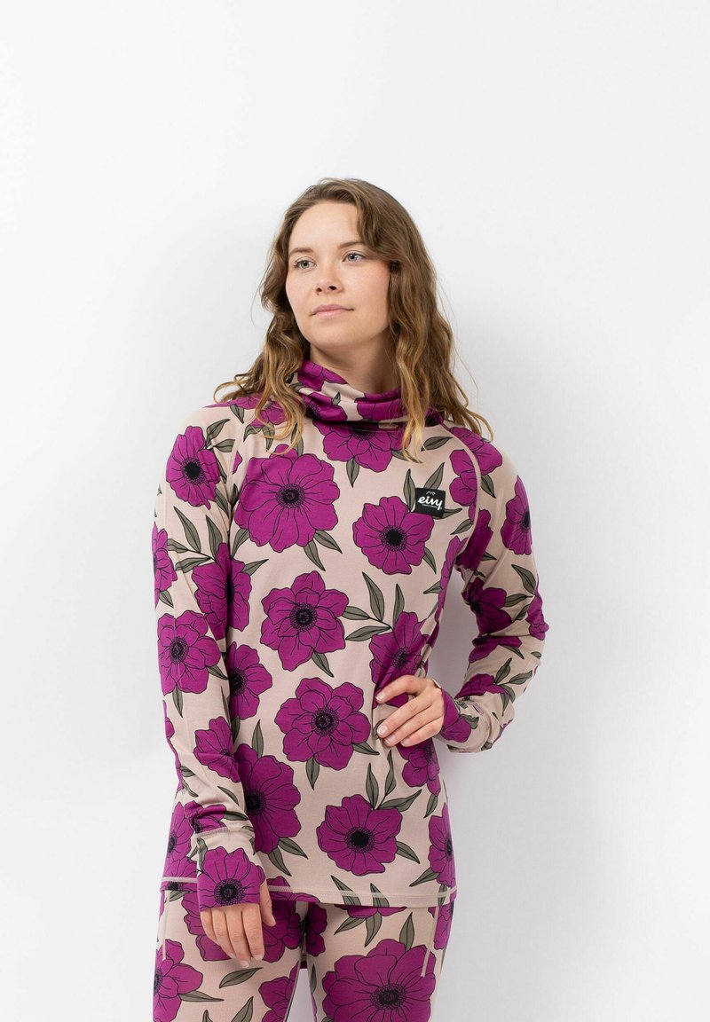 Beige Langarmoberteil mit großem lila Blumenmuster und grünen Blättern. Verfügt über einen hohen Kragen und eng anliegende Ärmel. Markenlogo auf der Vorderseite.