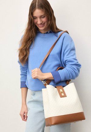 Kremo spalvos kibiro formos rankinė su rudos odos detalėmis, auksine sagtimi, geometriniu raštu ir ilgu dirželiu, derinama su mėlynu megztiniu ir dryžuotomis rankogaliais.