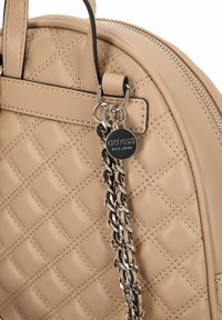 Bolso beige acolchado de cuero liso, con cierre de cremallera, tirante de cadena y una etiqueta metálica con la marca "GUESS" en relieve.