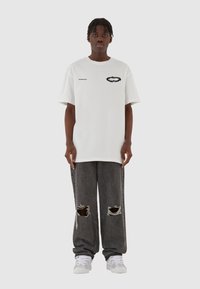 Hvid bomulds t-shirt med et sort grafisk design og logo, parret med løstsiddende grå slidte jeans og hvide sneakers.