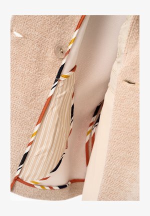 Beige textured blazer with striped inner pocket and multicolored trim, fastened with a clear button on a cream background.