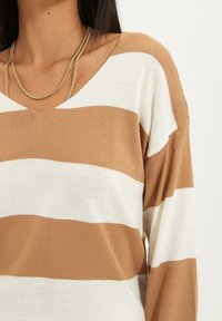 Gestreepte trui met bruin en witte horizontale lijnen, V-hals ontwerp, textuur gebreid materiaal, voorzien van een gelaagde gouden ketting.