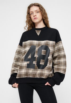 Jeune femme aux cheveux ondulés portant un sweat à carreaux noir et beige avec le numéro 48, debout devant un fond blanc uni.