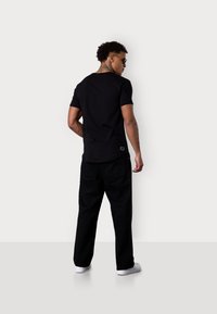 Schwarzes T-Shirt mit kurzen Ärmeln und geradem Saum, kombiniert mit lockeren schwarzen Hosen. Verfügt über ein Logoteil am hinteren Saum des Shirts. Weiße Sneakers.