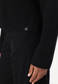 Maglione nero a coste con un design corto. Un piccolo etichetta nera con "cK" è visibile all'orlo. Indossato con jeans neri.