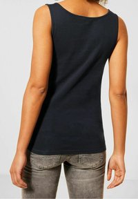 Svart ärmlös topp, mjukt tyg, rund halslinning, figurnära design. Bärs med grå jeans med subtila sömmar på fickorna.