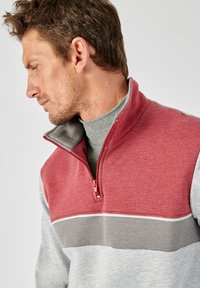 Sudadera de media cremallera en tela roja, gris y gris claro. Presenta un cuello alto y una textura suave con un diseño a bloques de color.
