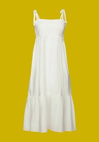 Robe maxi blanche sans manches avec encolure carrée, corsage en dentelle, bretelles nouées aux épaules et jupe à volants froncée sur fond jaune.
