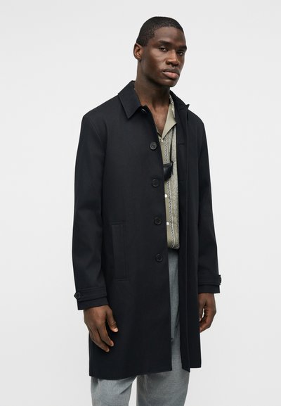 Άνδρας φορώντας μαύρο παλτό με κουμπιά, πουκάμισο με σχέδια, γκρι παντελόνι, στέκεται μπροστά σε απλό ανοιχτόφωτο φόντο με ουδέτερη έκφραση.