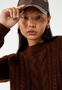 Pull en maille torsadée marron avec un col montant, associé à une casquette marron foncé. Texture lisse et motif de tressage bien visible.