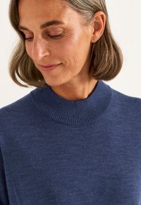 Kvinna med kort brunt hår som tittar ner, iklädd en blå ribbad rundhalsad tröja mot en ljus enkel bakgrund.