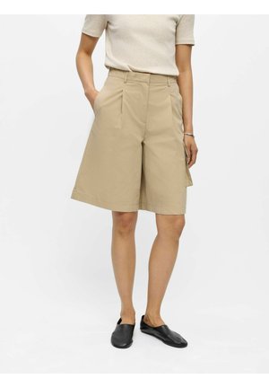 Persoon draagt beige wijdvallende knielange korte broek, lichtbeige top met korte mouwen, zwarte instapschoenen en een zilveren armband, staand met handen in de zakken.