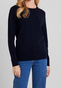 Tmavě modrý svetr s kulatým výstřihem a dlouhými rukávy, vyrobený z měkké látky. Má malou logo na hrudi a žebrované manžety.