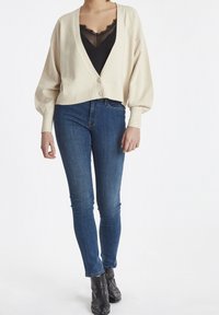 Elävävalkoinen, paksu neule cropped-neulejakku syvällä V-kaula-aukolla ja kahdella napilla; päällä musta läpikuultava pitsitoppi ja siniset kapeat farkut.