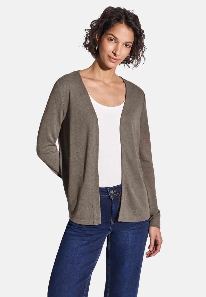 Frau mit kurzen lockigen Haaren trägt eine taupefarbene Strickjacke mit offenem Vorderteil über einem weißen Top mit Rundhalsausschnitt und dunklen blauen Jeans, steht vor einem schlichten Hintergrund.