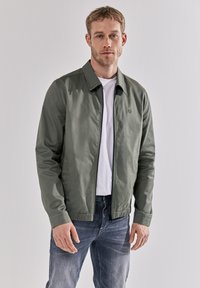 Světle zelená bomber bunda s límečkem, zipem a elastickými manžetami, nošená přes bílý tričko a modré džíny.