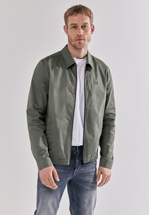 Une veste bomber vert clair avec un col, une fermeture éclair et des poignets élastiques, portée sur un t-shirt blanc et un jean bleu.