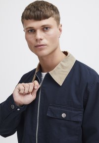 Marinblå jacka med beige krage, frontdragkedja och bröstficka. Har stickade detaljer och metallknappar. Tillverkad av bomull.