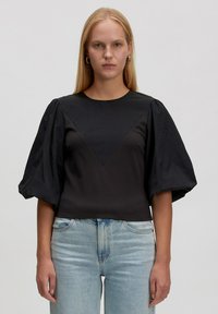Vrouw met lang blond haar, gekleed in een zwart shirt met pofmouwen en lichtblauwe spijkerbroek, staand tegen een effen achtergrond.