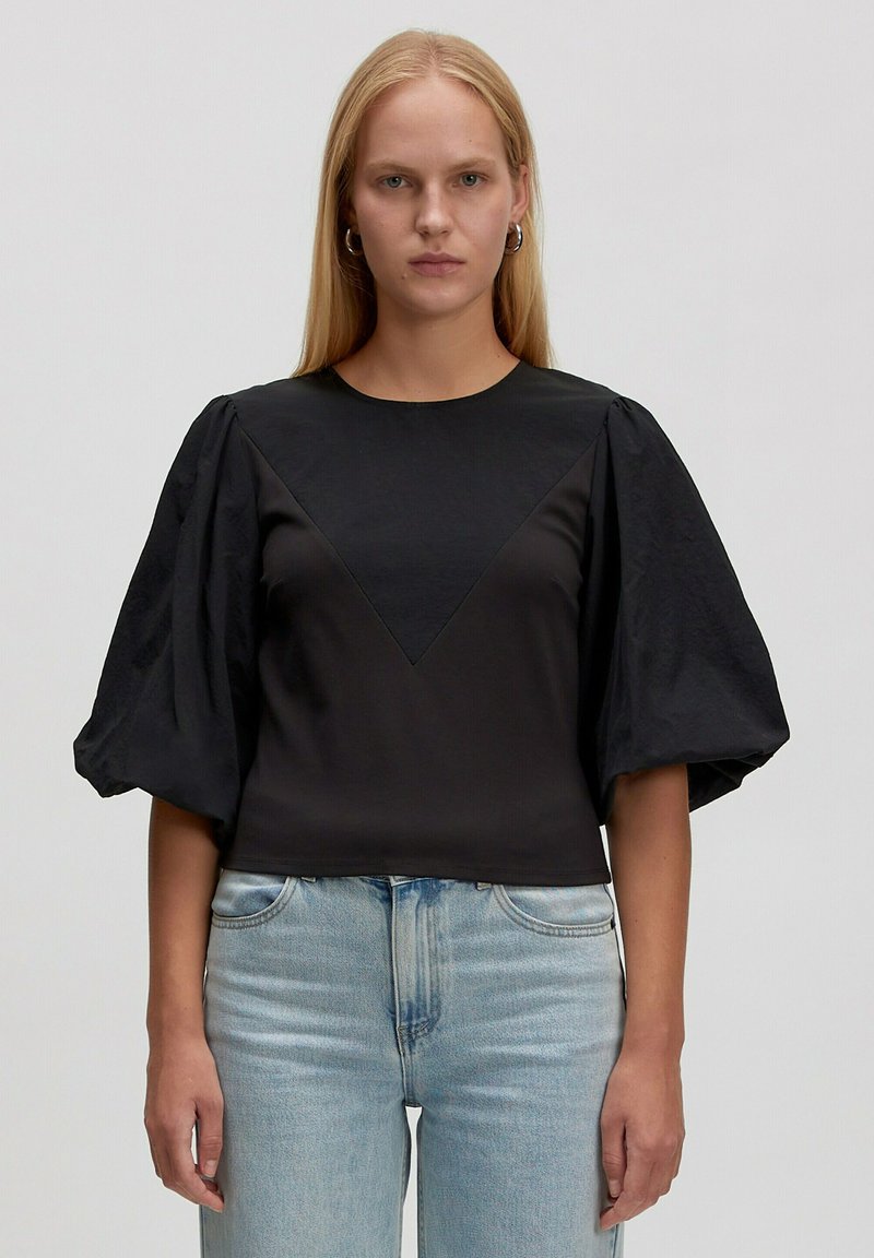 Vrouw met lang blond haar, gekleed in een zwart shirt met pofmouwen en lichtblauwe spijkerbroek, staand tegen een effen achtergrond.