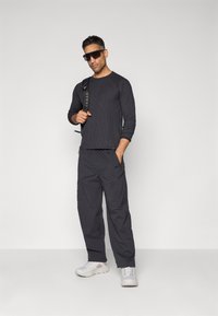 Schwarzes langärmliges geripptes Oberteil, kombiniert mit dunklen Cargo-Hosen. Graue Sneakers und dunkle Sonnenbrille runden den Look ab. Robuster Outdoor-Stil.