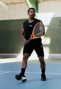 Bărbat îmbrăcat într-un tricou negru fără mâneci și pantaloni scurți ține o rachetă de tenis portocalie pe un teren de tenis albastru, evidențiindu-și constituția atletică și încălțămintea.