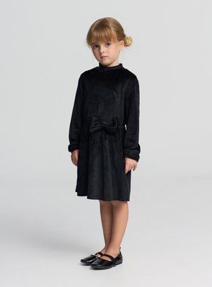 Czarna sukienka ze sztruksu z długimi rękawami, okrągłym dekoltem i ozdobną kokardą z przodu. Dopełnieniem stroju jest para czarnych lakierowanych butów.