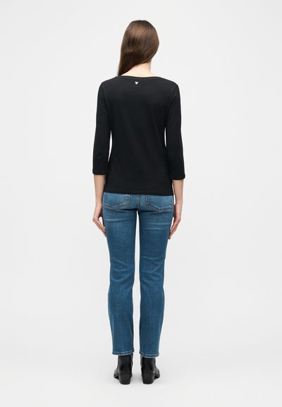 Γυναίκα με μακριά καστανά μαλλιά που φοράει μαύρο τοπ με μανίκια μέχρι τον αγκώνα, μπλε τζιν και μαύρα μποτάκια αστραγάλου, στέκεται γυρισμένη με την πλάτη.