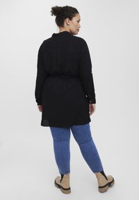 Vestido camisero negro de manga larga con cintura ceñida, presenta un collar y pliegues en la espalda, combinado con pantalones de mezclilla azul y botines beige.