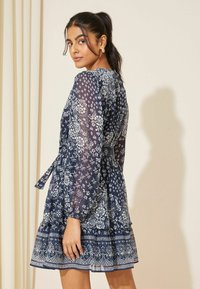 Rochie bleumarin cu mâneci lungi și bufante, cu un model floral. Dispune de o talie legată și margine cu volane stratificate, realizată dintr-un material ușor.