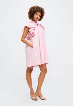 Γυναίκα με σγουρά μαλλιά που φορά ένα φαρδύ ροζ φόρεμα με βολάν στα μανίκια και μπεζ παπούτσια ανοιχτά στη μύτη, στέκεται με το ένα χέρι στην τσέπη.