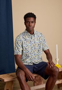 Korte mouw button-up shirt in een bloemenbladpatroon met blauwe, groene en gele kleuren, gemaakt van lichte stof; gedragen met marineblauwe shorts.