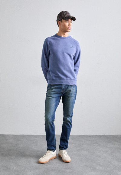 Kék crewneck pulóver kis logóval, kék farmernadrággal párosítva. Fehér sneakers, gumitalppal. Sötét tónusú csíkos sapka.