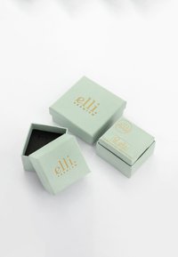 To lysgrønne gaveæsker med guldskrift, hvor den ene er åben og afslører en sort inderside. Æskerne har firkantede former og minimalistiske designelementer.