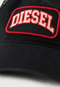 Melna cepure ar acs aizmuguri, kurai ir izteikta izšūta "DIESEL" logo sarkānā un krēmkrāsā uz plāksnes. Gluda auduma tekstūra, izliekts malums.