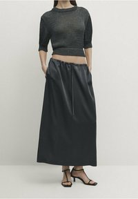 Haut noir à manches courtes en maille, associé à une longue jupe en satin foncé avec poches latérales et taille à cordon. Sandales noires à lanières complètent la tenue.