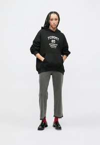 Μαύρο φούτερ με κουκούλα με την επιγραφή «TOMMY 85» στο μπροστινό μέρος, συνδυασμένο με γκρι τζιν, κόκκινες κάλτσες και μαύρα παπούτσια. Το φούτερ έχει μπροστινή τσέπη.