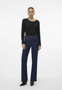Melna garām piedurknēm tops, kas kombinēts ar augstvērtīgiem, izplešamiem tumšo džinsu biksēm. Modelis stāv melnās papēžu zābakos. Vienkāršs, tīrs dizains.