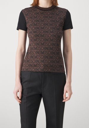 Kvinde iført en tætsiddende brun og sort top med korte ærmer med gentaget "MOSCHINO" tekstmønster, kombineret med sorte skræddersyede bukser.