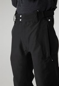 Personne portant un pantalon noir taille haute avec deux boutons argentés, une sangle latérale réglable, une poche zippée et un haut gris foncé.