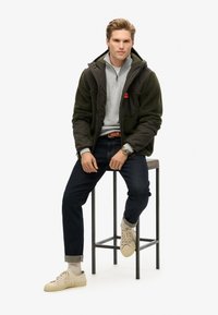 Giacca di pile verde oliva sopra una felpa grigia, jeans scuri e sneakers beige, seduto su uno sgabello, con un orologio al polso sinistro.