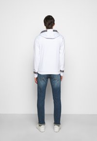 Felpa bianca con cappuccio, caratterizzata da un motivo logo nero sul colletto. Abbinata a jeans in denim blu e sneakers bianche.