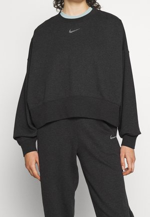 Persoon die een donkergrijze Nike-trui en bijpassende joggingbroek draagt met zichtbare Nike-logo's op de borst en dij.