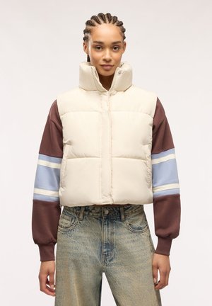 Giovane con i capelli intrecciati che indossa un gilet imbottito color crema sopra una felpa a righe marrone e blu e jeans azzurri.