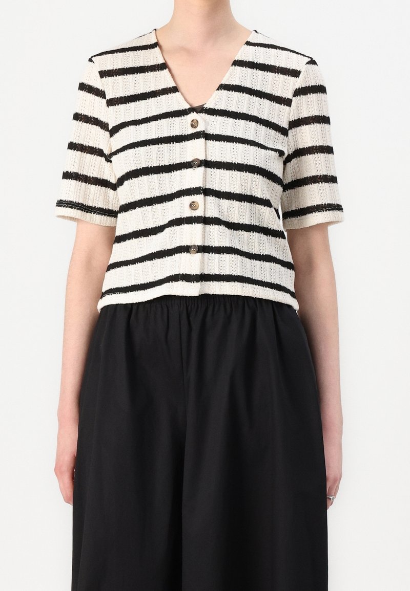 Femme portant un haut en maille blanc à manches courtes avec des rayures horizontales noires et des boutons, associé à un pantalon noir ample.