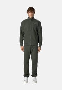 Giacca zip-up verde scuro con colletto alto e logo; pantaloni jogger abbinati; entrambi realizzati in morbido tessuto; chi la indossa ha scarpe da ginnastica bianche.