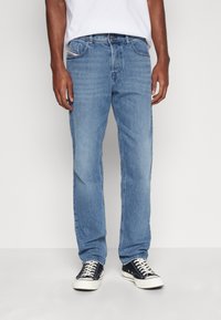 Vaqueros de mezclilla azul claro, corte recto, cinco bolsillos, detalles de costura, aspecto desgastado, combinados con zapatillas negras, sobre un fondo neutro.