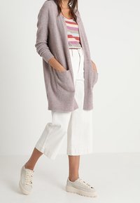 Cardigan en tricot violet clair avec devant ouvert, poches latérales, porté sur un débardeur à rayures, associé à un pantalon blanc large et court et des baskets beiges.